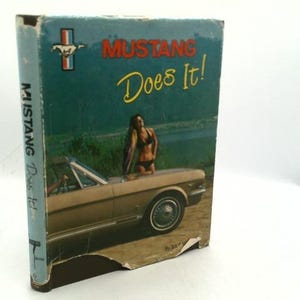 May include: A vintage hardcover book titled Mustang Does It! featuring a light blue cover with a tan convertible car, a person in a black bikini standing by the vehicle, the Ford Mustang logo, and visible wear along the bottom edge of the spine.