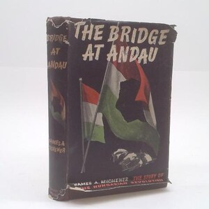 May include: A hardcover book titled "The Bridge at Andau" with a dark cover featuring the Hungarian flag. The book's title and author's name are visible. The book's spine also displays the title. The book is set against a white background.
