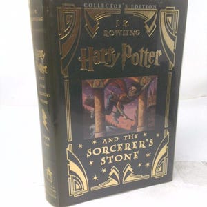 May include: A black hardcover book titled Harry Potter and the Sorcerer's Stone, Collector's Edition by J.K. Rowling. The cover features gold foil art deco accents and a central illustration of a character on a broomstick against a purple sky.