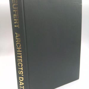 May include: A black hardcover book titled NEUFERT ARCHITECTS' DATA printed in gold lettering on the spine. The book is positioned vertically, showing the front cover and the spine, which features a small gold circular logo at the bottom.