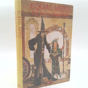 May include: A hardcover book titled Equal Rites by Terry Pratchett with a tan spine and a cover illustration featuring a tall figure in a black robe and pointed hat standing next to a child holding a staff and a book, with a black cat nearby.