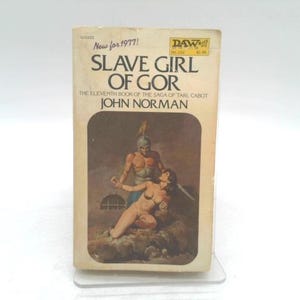 May include: A vintage paperback book titled "Slave Girl of Gor" by John Norman. The cover art depicts a muscular man and a woman in a fantasy setting. The book is labeled "New for 1977!" and has a "DAW" logo.