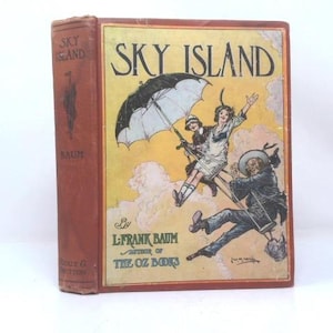 May include: A hardcover book titled Sky Island by L. Frank Baum with a red spine and a front cover illustration featuring two figures suspended from a large black umbrella against a yellow and pink sky. The text reads Sky Island and The Oz Books.
