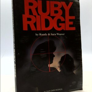 May include: A black and white book cover with the title "The Federal Siege at Ruby Ridge" in red letters. The cover also features a silhouette of a man and a child in a crosshairs.