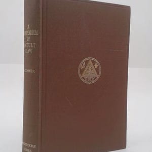 May include: A brown hardcover book titled "A Compendium of Occult Law" by Clymer. The book has a gold emblem on the front cover and text on the spine. The book is part of the Rosicrucian Series.