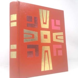 May include: A red book with gold and maroon geometric designs on the cover. The spine has gold rectangular accents. The cover design features a central square with vertical and horizontal gold shapes and maroon and pink accents.
