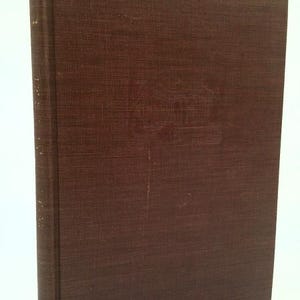 May include: A hardcover book with a brown cover. The cover has a textured appearance and a subtle embossed design. The spine is also brown and shows signs of wear. The book is slightly angled, showcasing its aged condition.
