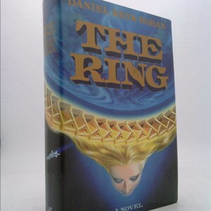 May include: A book cover with a blue and black background. The title "THE RING" is in large, bold, gold letters. The cover features a woman's face and hair, with a golden ring around the top of her head.