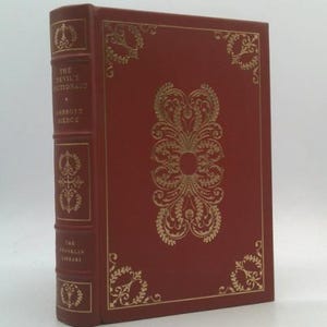 May include: A hardcover book titled "The Devil's Dictionary" by Ambrose Bierce. The book has a red cover with gold decorative accents and text. The spine also features gold text and embellishments. The Franklin Library edition.