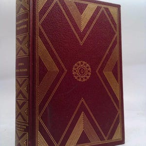 May include: A hardcover book titled "Manhattan Transfer" by John Dos Passos. The book has a burgundy cover with gold geometric designs and text. The spine also features text and the International Collectors Library logo.