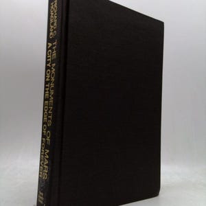 May include: A black hardcover book titled The Monuments of Mars: A City on the Edge of Forever by Richard C. Hoagland. The title and author name are printed in gold lettering on the spine of the book, which is positioned vertically.