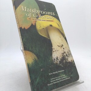 May include: A book titled "Mushrooms of Colorado and the Southern Rocky Mountains" is displayed. The cover features a large mushroom with a yellow cap and white stem against a green background. The book is propped up on a clear stand.