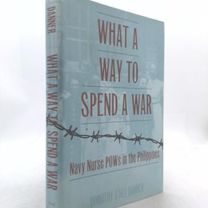 May include: A hardcover book titled What a Way to Spend a War: Navy Nurse POWs in the Philippines by Dorothy Still Danner. The light blue cover features a dark red title, a graphic of barbed wire, and a faded black and white photograph of nurses.