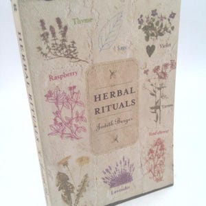 May include: A hardcover book titled "HERBAL RITUALS" by Judith Berger. The cover is beige with illustrations of herbs like thyme, raspberry, sage, violet, yarrow, red clover, dandelion, and lavender. The book has a textured, aged appearance.