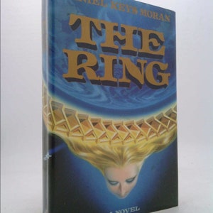 May include: A book cover with a blue and gold design. The title "THE RING" is in large, bold, black letters. The author's name, "DANIEL KEYS MORAN", is printed in smaller letters above the title. The cover features a woman's face and hair, with a gold ring above her head.