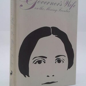 May include: A hardcover book titled A Governor's Wife on the Mining Frontier with a light gray cover. The front features a large, stylized black graphic illustration of a face. The title is printed in a dark purple script font at the top.