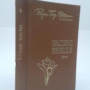 May include: A brown book titled "Pacific Shells" by Morris, part of the Roger Tory Peterson Field Guides series. The title and author's name are in gold lettering. A seashell illustration is also in gold.