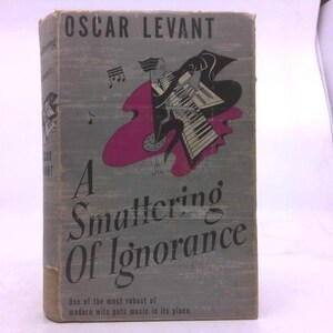 May include: A vintage book titled "A Smattering of Ignorance" by Oscar Levant. The cover is gray with a pink and black illustration of a musician playing a piano. The text on the cover is in a serif font.
