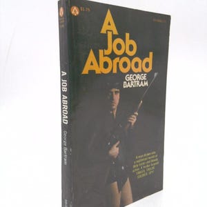 May include: Vintage paperback book titled "A Job Abroad" by George Bartram. The cover features a man in a dark coat holding a weapon. The title is in large yellow letters against a black background. The book is a thriller.