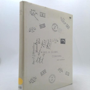 May include: A vintage book with a cream-colored cover, featuring postal stamps and handwritten text. The cover includes the text "GEO. H. SILSBY, CONCORD" and various postmarks dated from 1902 to 1905. The book has a yellow sticker with "RB" on the spine.