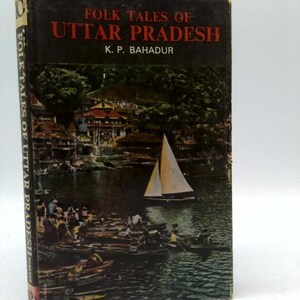 May include: A vintage book titled "Folk Tales of Uttar Pradesh" by K. P. Bahadur. The cover features a scenic illustration of a lake with boats, a sailboat, and buildings. The book has a dark cover with red and white text.
