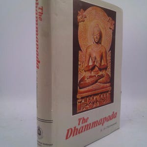 May include: A hardcover book titled "The Dhammapada" with a beige cover. The cover features a reddish-brown image of a seated Buddha figure. The title is in red, and the spine also has the title printed vertically.