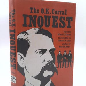 May include: A red book cover with the title "The O.K. Corral INQUEST" in black text. The cover features a black and white illustration of a man with a mustache and a group of men in hats.