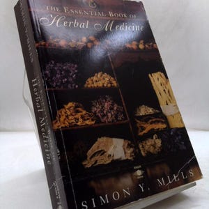 May include: A hardcover book titled The Essential Book of Herbal Medicine by Simon Y. Mills. The dark brown cover features a grid of photographs displaying various dried herbs, roots, and botanical specimens arranged on dark wooden shelves.