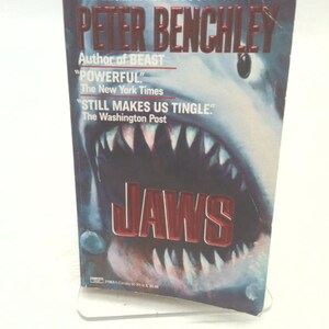 May include: A paperback book titled "JAWS" by Peter Benchley. The cover features a menacing shark with open jaws, set against a dark blue background. Quotes from The New York Times and The Washington Post are also visible.