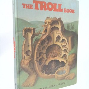 May include: A hardcover book titled "The Troll Book" by Michael Berenstain. The cover features a detailed illustration of a troll-shaped structure with internal mechanisms and figures. The background is a light blue sky and green landscape.