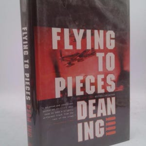 May include: A hardcover book titled "Flying to Pieces" by Dean Ing. The cover features a red background with the title in large white letters and an image of a plane. The author's name is also in white.
