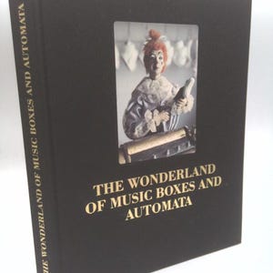 May include: A black hardcover book titled The Wonderland of Music Boxes and Automata with gold lettering on the spine and front cover. The front cover features a rectangular inset photograph of a clown figure with orange hair and a ruffled collar.