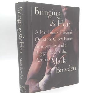 May include: A hardcover book titled Bringing the Heat by Mark Bowden. The black dust jacket features white text and a dark, shadowed photograph of a football player holding a football, with the title and author name printed on the spine.
