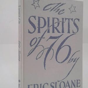 May include: A hardcover book titled "The Spirits of '76" by Eric Sloane. The book has a light gray cover with blue lettering and star illustrations. The book is standing upright.