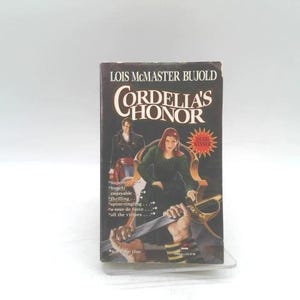 May include: A hardcover book titled "Cordelia's Honor" by Lois McMaster Bujold. The cover features a woman in a green dress holding a sword, with a man in a black coat in the background. The book has a red "Best Seller" sticker.