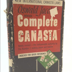 May include: A black and red book titled "Oswald Jacoby's Complete Canasta" with the words "New International Canasta Laws" and "Revised" on the cover. The book is indexed for quick reference and features a green table with playing cards on it.