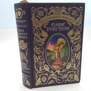 May include: A hardcover book titled "Classic Fairy Tales" by Hans Christian Andersen. The book has a dark purple cover with gold floral and scroll designs. The cover features an oval illustration of a fairy holding a flower. The spine has the author's name.