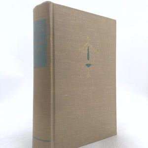 May include: A hardcover book with a tan cloth cover featuring a gold-stamped emblem on the front. The spine has a dark teal label with gold text. The book is standing upright, showing the front cover and the spine against a plain white background.