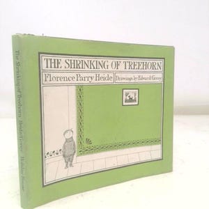 May include: A hardcover book titled "THE SHRINKING OF TREEHORN" by Florence Parry Heide, with drawings by Edward Gorey. The cover is green with a framed illustration of a child and a green wall. The book's spine also has the title and author's name.