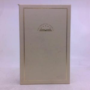 May include: A rectangular off-white book slipcase featuring a thin gold border line. Centered near the top is a gold emblem depicting an open book with five stars arranged in a semi-circle above it, with the text THE LIBRARY OF AMERICA.