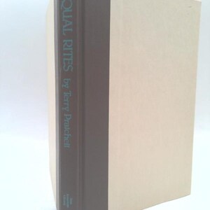 May include: A hardcover book titled Equal Rites by Terry Pratchett. The book features a dark brown spine with light blue text and cream-colored covers. The New American Library logo is visible at the bottom of the spine on this upright volume.
