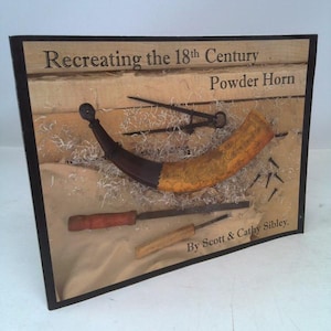 May include: A display featuring a powder horn and tools, with the text "Recreating the 18th Century Powder Horn" and "By Scott & Cathy Sibley." The horn is tan and brown, with a dark handle. Tools and nails are also displayed.