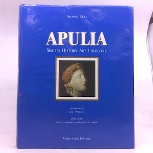 May include: A blue hardcover book titled APULIA Sights History Art Folklore by Stefania Mula. The cover features a central square image of a classical stone bust with gold earrings, with text credits for Lino Patruno, Nicola Amato, Sergio Leonardi, and Mario Adda Editore.