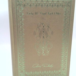 May include: A gold-colored book with a green border and the title "Lucky We Found Each Other" on the front cover. The author's name, "Gerry Farrell", is written in cursive below the title.