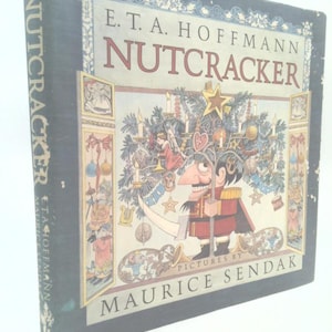 May include: A hardcover book titled Nutcracker by E.T.A. Hoffmann with illustrations by Maurice Sendak. The dark blue cover features a central illustration of a nutcracker character standing under a decorated tree with a gold star on top.