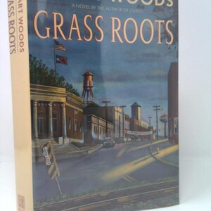 May include: A hardcover book titled "Grass Roots" by Stuart Woods. The cover features a town scene with buildings, a water tower, and an American flag. The book's spine also displays the title and author's name.