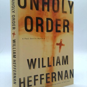 May include: A hardcover book titled "UNHOLY ORDER" by William Heffernan. The book has a tan cover with the title in large black letters and the author's name below. A cross graphic is visible. The book is a Paul Devlin Mystery.