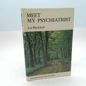 May include: A hardcover book titled "MEET MY PSYCHIATRIST" by Les Blacklock. The book has a cream-colored cover with black text and a green and brown image of a forest road. The book's spine reads "Blacklock".