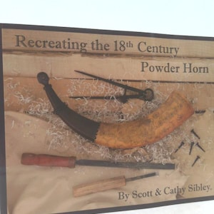 May include: A framed display featuring a powder horn and tools, with the text "Recreating the 18th Century Powder Horn" and "By Scott & Cathy Sibley." The horn is tan and black, with a curved shape. Tools include a red-handled tool and a wooden-handled tool.