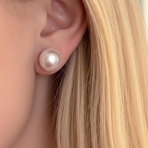 May include: A close-up of a large, round, pearl stud earring. The earring is a creamy white color and is set against the skin and blonde hair. The earring is a classic jewelry piece.